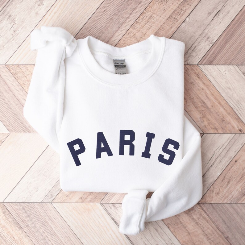 Puede incluir: Sudadera blanca con un gr&aacute;fico "PARIS" azul marino en la parte delantera.