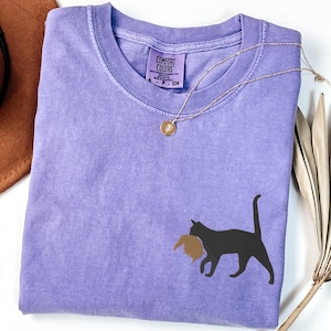 May include: A lavender colored t-shirt with a black silhouette of a cat carrying a brown bird in its mouth. The shirt has a tag that says "Comfort Colors" and "S - P - CH".