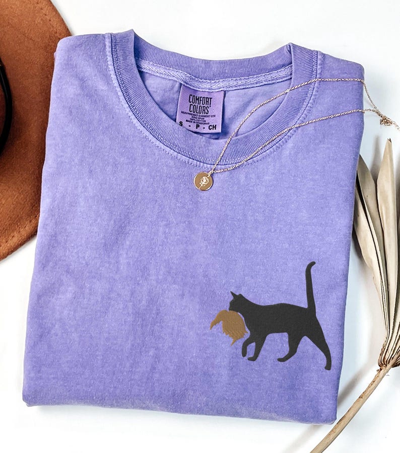 May include: A lavender colored t-shirt with a black silhouette of a cat carrying a brown bird in its mouth. The shirt has a tag that says "Comfort Colors" and "S - P - CH".