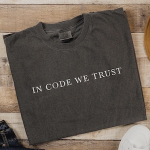 May include: A dark gray t-shirt with the text "IN CODE WE TRUST" printed in white on the front.