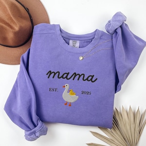 May include: A lilac-colored sweatshirt with the word "mama" embroidered in black script. Below the word "mama" is an embroidered image of a goose and a chick. The text "EST. 2025" is below the goose and chick. A gold necklace is draped on the sweatshirt.