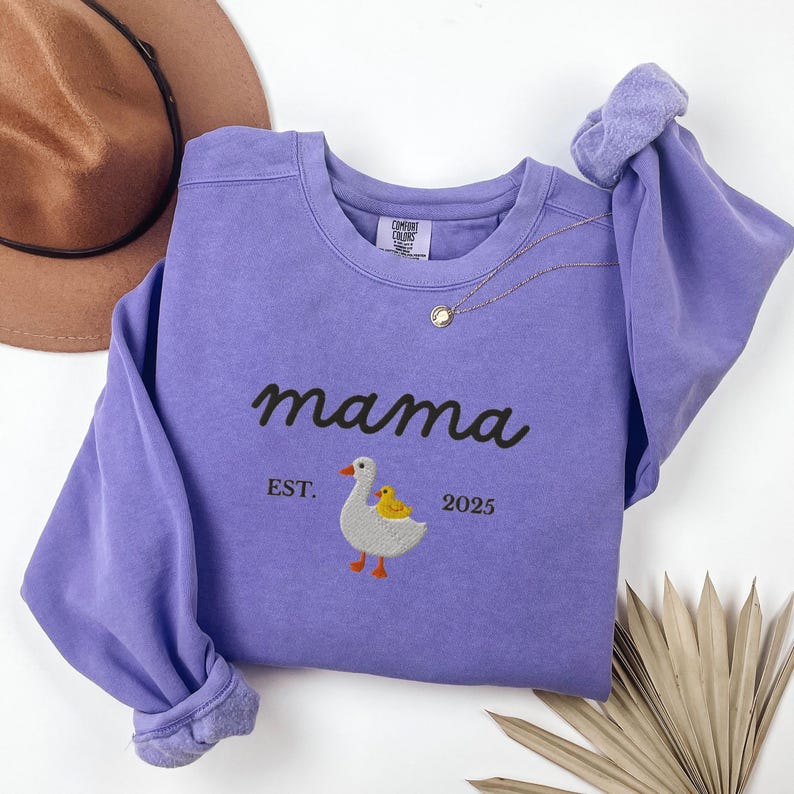 May include: A lilac-colored sweatshirt with the word "mama" embroidered in black script. Below the word "mama" is an embroidered image of a goose and a chick. The text "EST. 2025" is below the goose and chick. A gold necklace is draped on the sweatshirt.