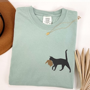 May include: A light blue t-shirt with a black cat silhouette carrying a brown bird in its mouth. The shirt has a tag that says "Comfort Colors" on it.