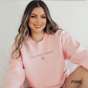 May include: A light pink sweatshirt with the text "TILE WHISPERER" in black. Below the text is a small graphic. The word "PINK" is printed in the bottom right corner. The sweatshirt is a solid color.