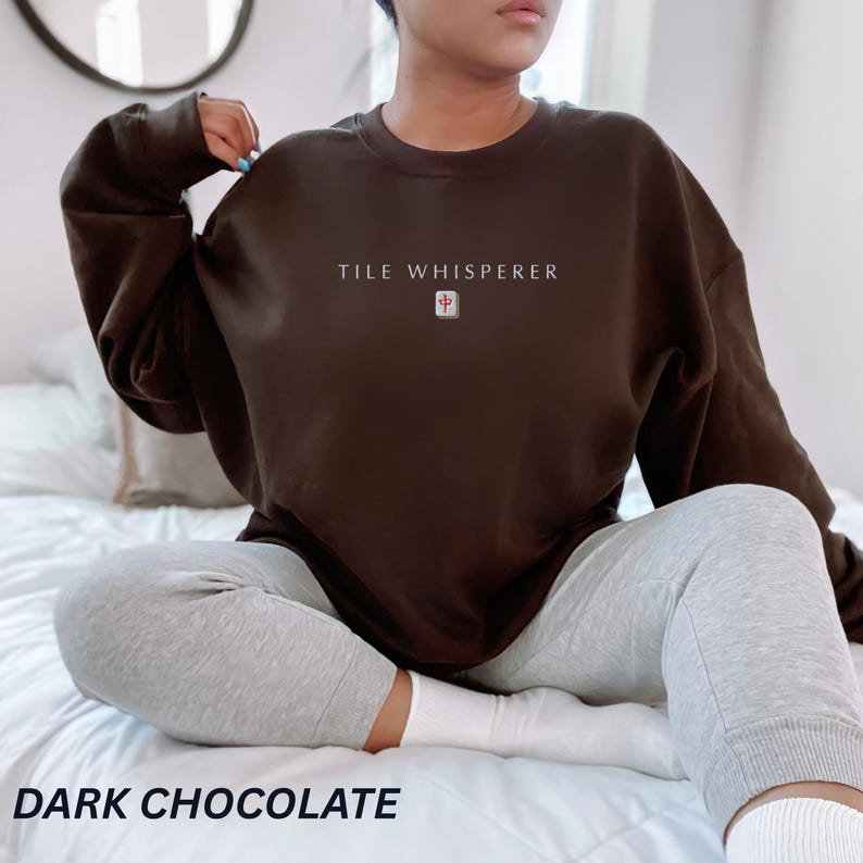 May include: Dark chocolate brown sweatshirt with the text "TILE WHISPERER" and a small tile graphic. The sweatshirt is paired with gray sweatpants. The words "DARK CHOCOLATE" are at the bottom of the image.