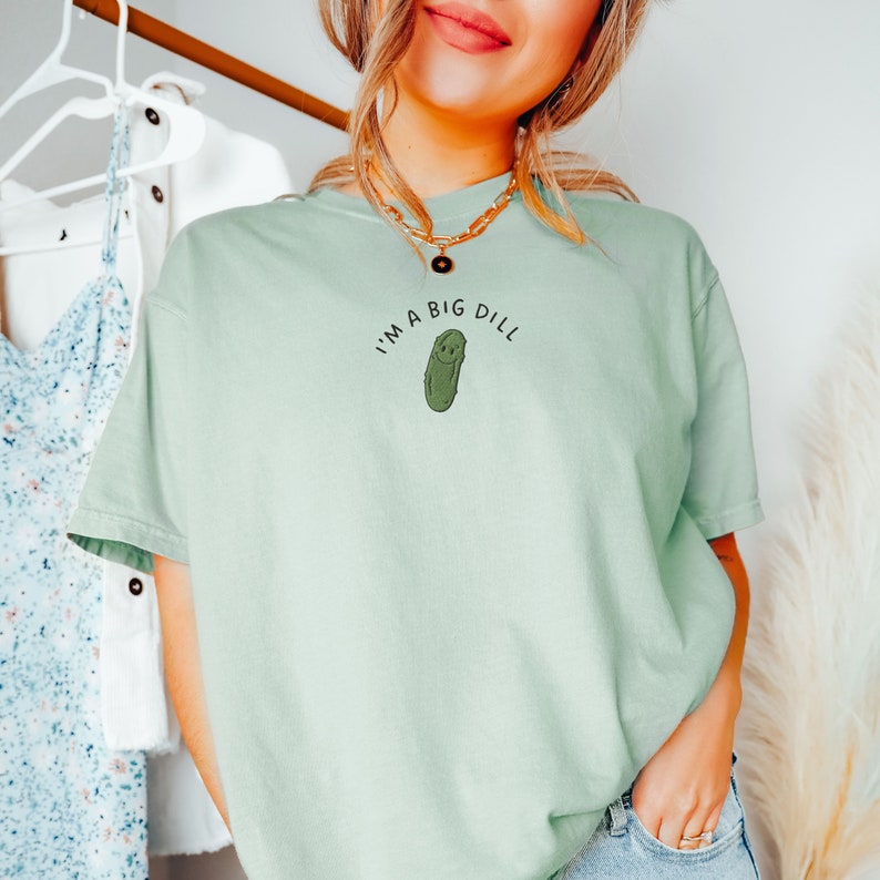 Op de afbeelding: Een lichtgroen T-shirt met de tekst "I'm a big dill" en een cartoon-komkommer met een glimlach.