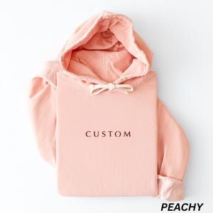 May include: A peach-colored hoodie with a drawstring hood and a front pocket. The word "CUSTOM" is printed in a dark brown font on the front. The word "PEACHY" is printed in a black font at the bottom.