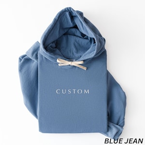 May include: A blue jean colored hoodie with a drawstring hood. The word "CUSTOM" is printed in white on the front. The bottom right corner of the image has the words "BLUE JEAN" printed in black.