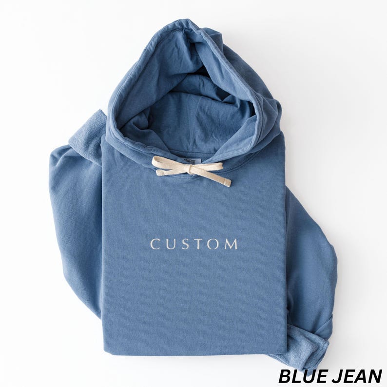 May include: A blue jean colored hoodie with a drawstring hood. The word "CUSTOM" is printed in white on the front. The bottom right corner of the image has the words "BLUE JEAN" printed in black.
