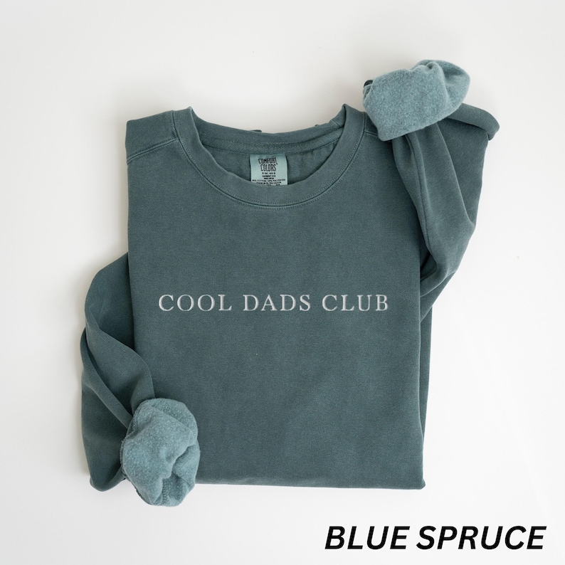 May include: Blue spruce sweatshirt with the text "COOL DADS CLUB" in white. The sweatshirt has a crew neck and long sleeves with folded cuffs. The bottom of the image has the text "BLUE SPRUCE" in a bold, black font.