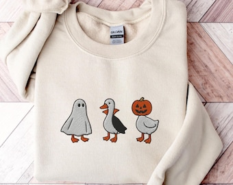 May include: Cream-colored sweatshirt featuring embroidered Halloween-themed characters: a ghost, a duck with black wings, and a duck with a jack-o'-lantern head. The sweatshirt has a crew neck and long sleeves. The brand tag is visible on the inside of the collar.
