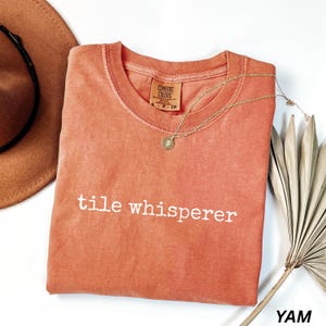 K&ouml;nnte beinhalten: Korallfarbenes T-Shirt mit dem wei&szlig;en Text "tile whisperer". Eine goldene Halskette mit Anh&auml;nger liegt auf dem Shirt. Ein brauner Hut und ein getrocknetes Palmblatt sind ebenfalls im Bild.
