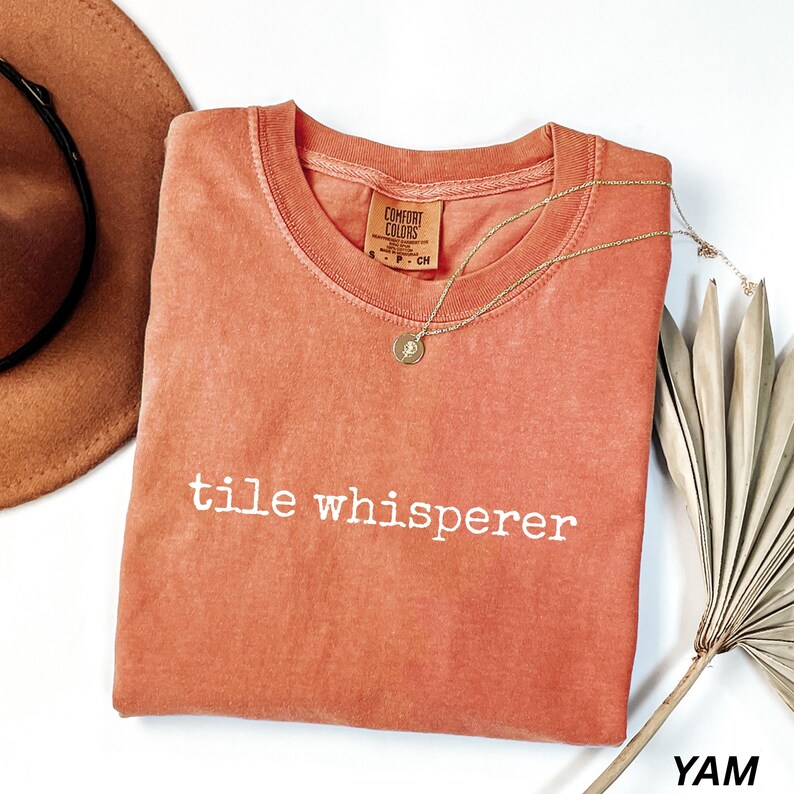 K&ouml;nnte beinhalten: Korallfarbenes T-Shirt mit dem wei&szlig;en Text "tile whisperer". Eine goldene Halskette mit Anh&auml;nger liegt auf dem Shirt. Ein brauner Hut und ein getrocknetes Palmblatt sind ebenfalls im Bild.