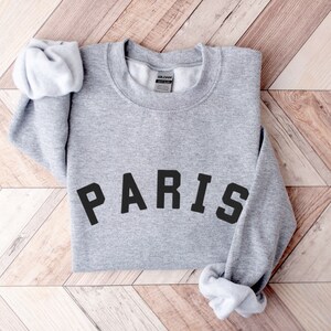 Puede incluir: Sudadera gris con la palabra "PARIS" impresa en letras negras.
