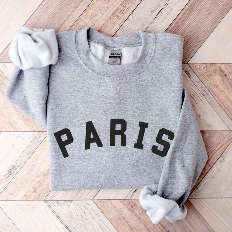Puede incluir: Sudadera gris con la palabra "PARIS" impresa en letras negras.