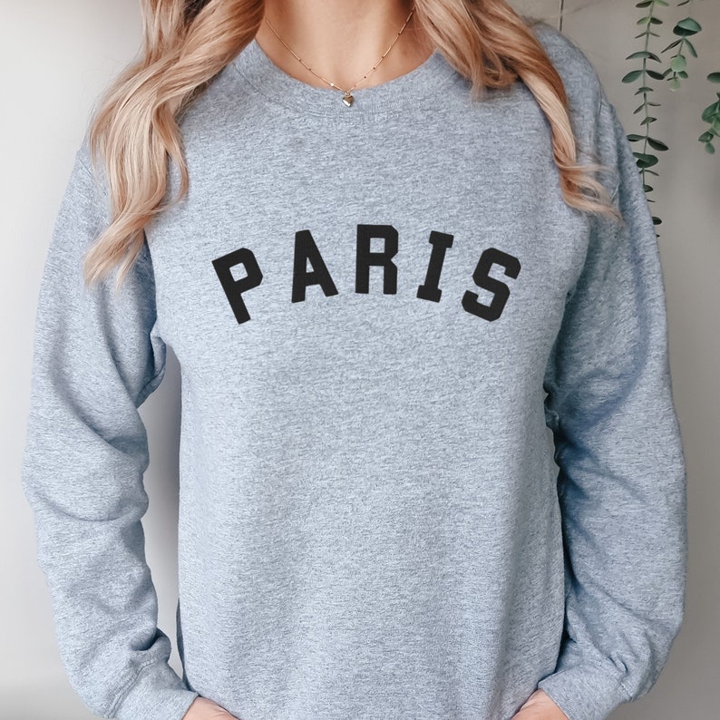 Puede incluir: Una sudadera gris con la palabra "PARIS" impresa en negro en la parte delantera.