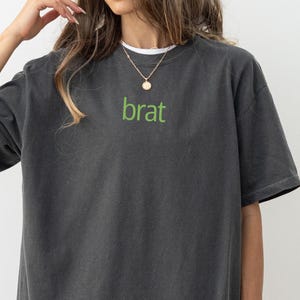 May include: A dark gray t-shirt with the word "brat" printed in green on the front. The shirt has a relaxed fit and a crew neck.