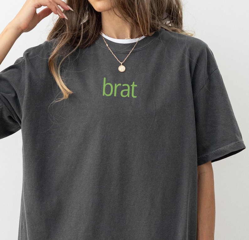 May include: A dark gray t-shirt with the word "brat" printed in green on the front. The shirt has a relaxed fit and a crew neck.