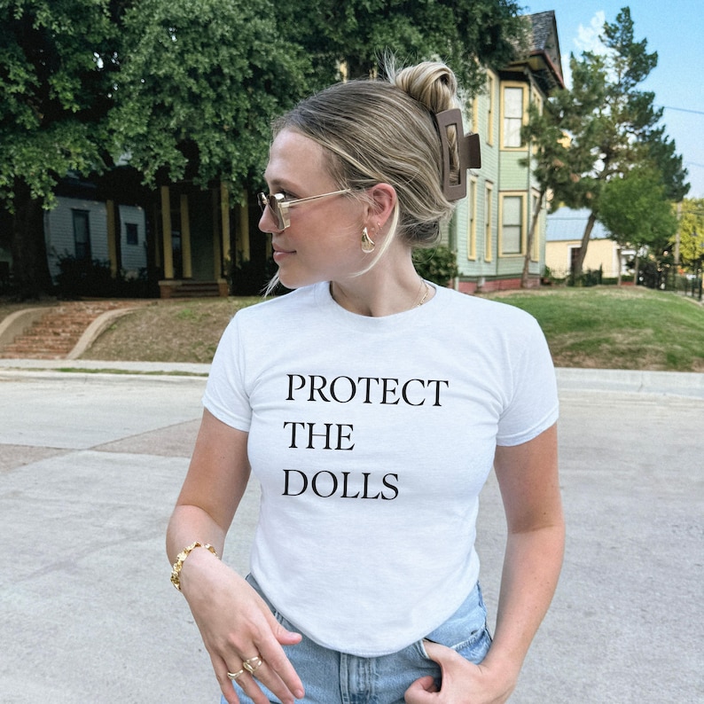May include: White t-shirt with the text "PROTECT THE DOLLS" in black. The person is wearing gold-framed sunglasses, gold earrings, and a gold bracelet. A brown hair clip holds up the hair.