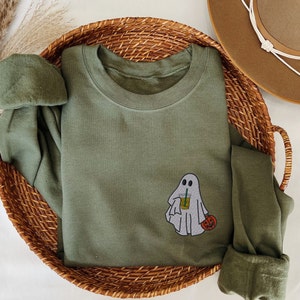 May include: A green long-sleeved shirt with a ghost holding a cup of iced coffee and a jack-o-lantern. The shirt is on a woven basket.