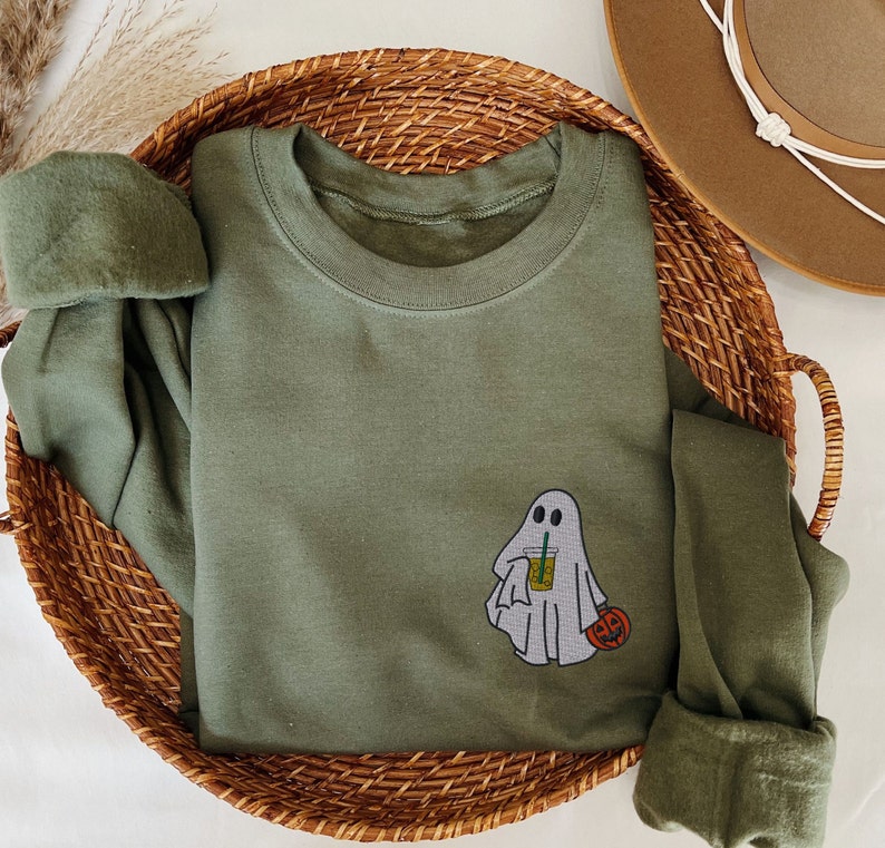 May include: A green long-sleeved shirt with a ghost holding a cup of iced coffee and a jack-o-lantern. The shirt is on a woven basket.