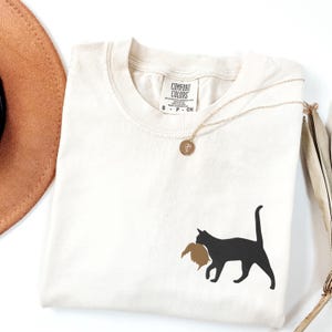 May include: A white t-shirt with a black silhouette of a cat carrying a brown bird in its mouth. The shirt has a tag that says "Comfort Colors" and "S - P - CH".