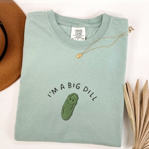 Op de afbeelding: Een lichtgroen T-shirt met de tekst "I'm a big dill" en een cartoon-komkommer met een smiley.