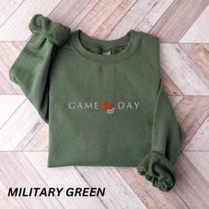 May include: Military green sweatshirt with the text "GAME DAY" and a basketball graphic. The sweatshirt is a crew neck style with long sleeves and ribbed cuffs. The text and graphic are in white and orange.