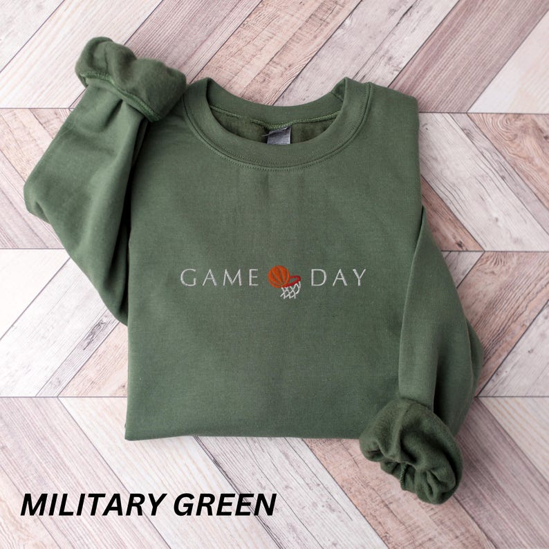 May include: Military green sweatshirt with the text "GAME DAY" and a basketball graphic. The sweatshirt is a crew neck style with long sleeves and ribbed cuffs. The text and graphic are in white and orange.