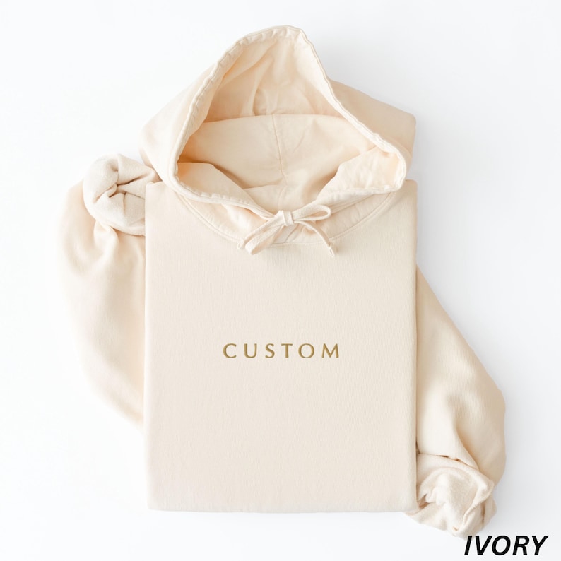 May include: Ivory-colored hoodie with the word "CUSTOM" printed in gold. The hoodie has a drawstring hood and long sleeves. The folded hoodie is on a white surface. The word "IVORY" is printed in the bottom right corner.