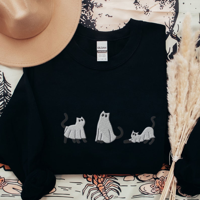 K&ouml;nnte beinhalten: Schwarzer Pullover mit einem wei&szlig;en gestickten Design von drei Katzen, die als Geister verkleidet sind. Der Pullover hat ein Gildan-Etikett unten links.