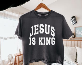 May include: A charcoal gray short-sleeve t-shirt featuring the white text JESUS IS KING in a bold, arched collegiate-style font across the chest. The garment is displayed on a wooden hanger against a neutral background, showcasing a crew neckline.