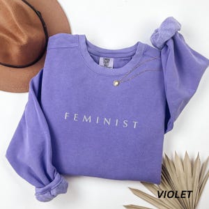 Pu&ograve; includere: Una felpa viola con la scritta "FEMINIST" stampata in bianco. Una collana d'oro con un ciondolo poggia sulla maglia. La manica &egrave; arrotolata. Un cappello marrone &egrave; visibile sullo sfondo.