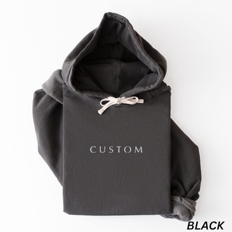May include: A black hoodie with a drawstring hood and the word "CUSTOM" printed in white. The word "BLACK" is printed in white at the bottom right corner. The hoodie is folded on a white surface.