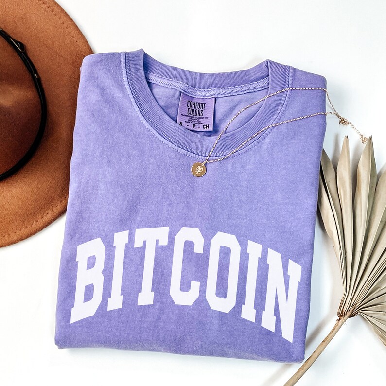 May include: A purple t-shirt with the word "BITCOIN" printed in white on the front. The shirt is folded and laid flat on a white surface. A brown hat and a dried palm leaf are also visible in the image.