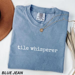 K&ouml;nnte beinhalten: Ein blaues Jeans-T-Shirt mit den wei&szlig;en Worten "tile whisperer". Das Shirt hat einen Rundhalsausschnitt und kurze &Auml;rmel. Eine goldene Halskette mit Anh&auml;nger ist &uuml;ber dem Shirt drapiert. Das Shirt ist mit "Comfort Colors" gekennzeichnet.
