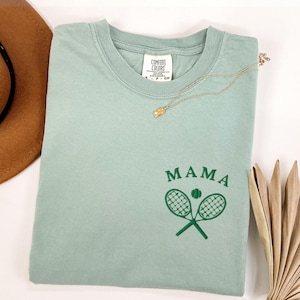 May include: A light green t-shirt with the text "MAMA" and a green tennis racket and ball graphic. The shirt is made by Comfort Colors.