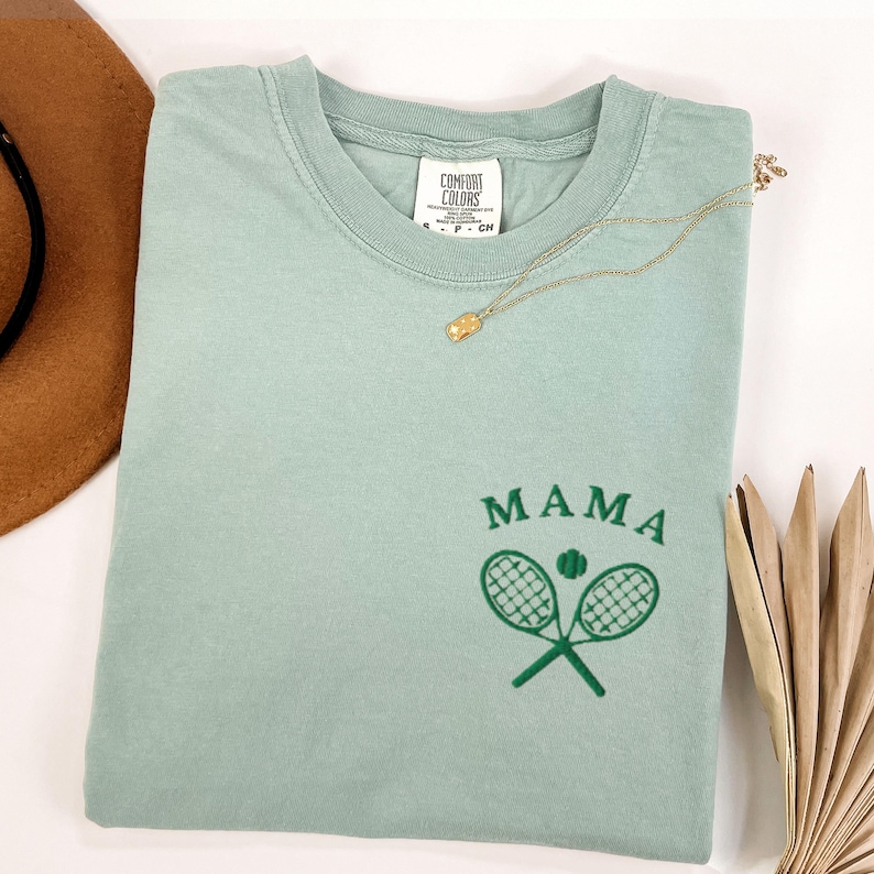 May include: A light green t-shirt with the text "MAMA" and a green tennis racket and ball graphic. The shirt is made by Comfort Colors.