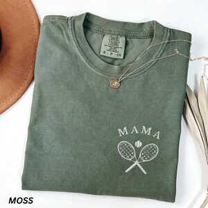 May include: Moss green t-shirt with the word "MAMA" above a tennis ball and crossed tennis rackets. A gold necklace with a pendant is on the shirt. The shirt has a Comfort Colors tag.