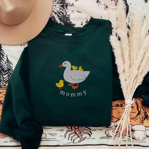 May include: A dark green sweatshirt with an embroidered image of a white goose with three yellow ducklings. The text "mommy" is embroidered below the goose.