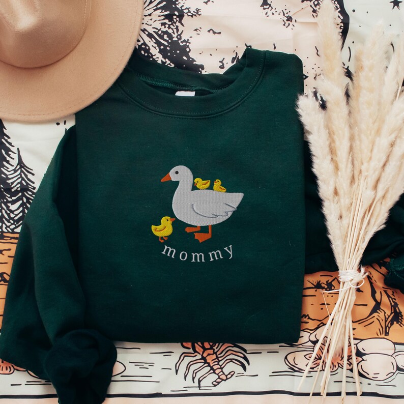 May include: A dark green sweatshirt with an embroidered image of a white goose with three yellow ducklings. The text "mommy" is embroidered below the goose.