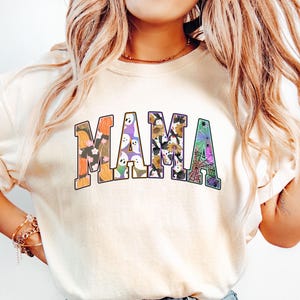 Puede incluir: Una camiseta blanca con la palabra "MAMA" impresa en una fuente colorida con tem&aacute;tica de Halloween. Las letras est&aacute;n llenas de im&aacute;genes de calabazas, fantasmas, ara&ntilde;as y otros motivos de Halloween.
