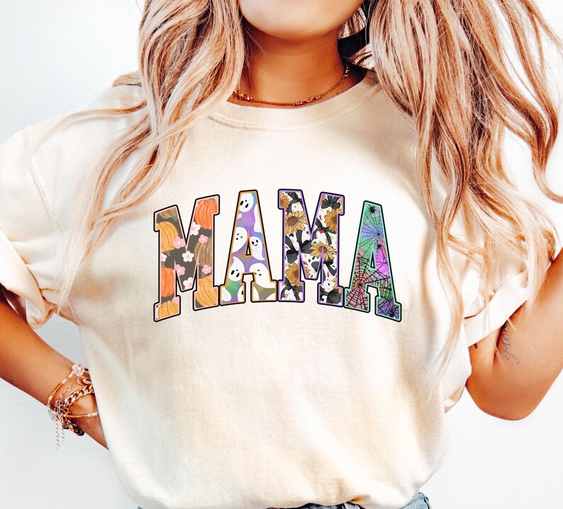 Puede incluir: Una camiseta blanca con la palabra "MAMA" impresa en una fuente colorida con tem&aacute;tica de Halloween. Las letras est&aacute;n llenas de im&aacute;genes de calabazas, fantasmas, ara&ntilde;as y otros motivos de Halloween.