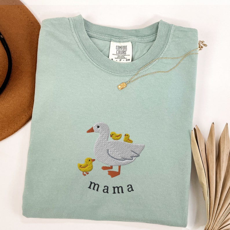 May include: A light blue t-shirt with an embroidered image of a goose and three goslings. The text "mama" is embroidered below the image.
