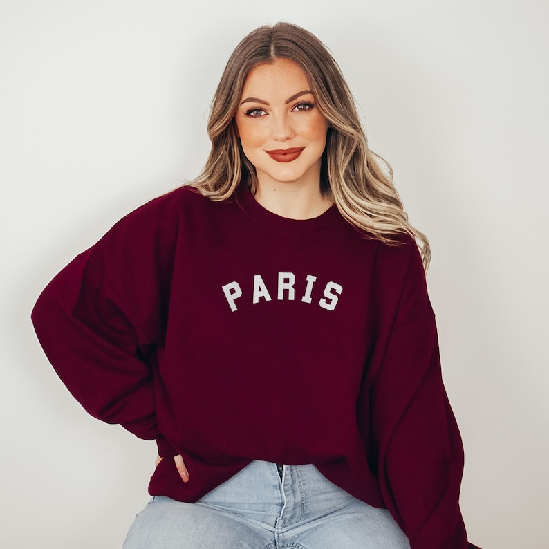 Puede incluir: Una sudadera color burdeos con la palabra "PARIS" impresa en blanco en la parte delantera. La sudadera la lleva puesta una persona con el pelo largo y rubio.