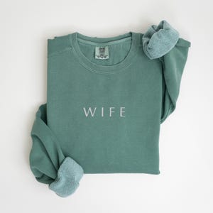 May include: A green sweatshirt with the word "WIFE" embroidered on the front. The sweatshirt is folded and the sleeves are rolled up.