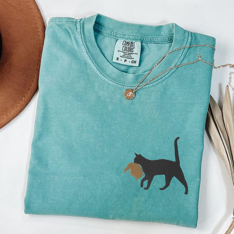 May include: A teal blue t-shirt with a black cat silhouette carrying a brown bird in its mouth. The shirt has a tag that reads "Comfort Colors" and "S - P - CH".