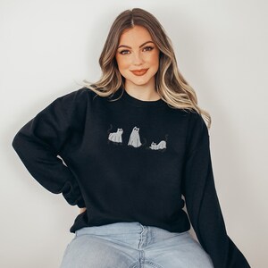 K&ouml;nnte beinhalten: Ein schwarzer Pullover mit einem Aufdruck von drei Geisterkatzen. Die Katzen sind wei&szlig; mit schwarzen Augen und Schw&auml;nzen. Der Pullover wird von einer Person mit langen blonden Haaren getragen.