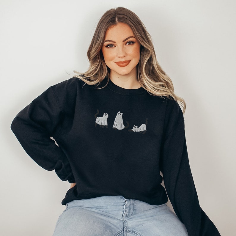 K&ouml;nnte beinhalten: Ein schwarzer Pullover mit einem Aufdruck von drei Geisterkatzen. Die Katzen sind wei&szlig; mit schwarzen Augen und Schw&auml;nzen. Der Pullover wird von einer Person mit langen blonden Haaren getragen.