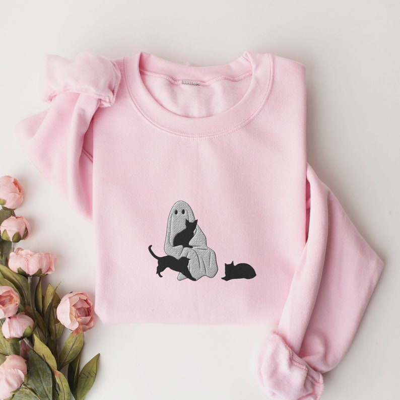 K&ouml;nnte beinhalten: Ein rosa Sweatshirt mit einem grauen Geist und drei schwarzen Katzen, die auf der Vorderseite gestickt sind. Der Geist h&auml;lt eine Katze und die anderen beiden Katzen sitzen auf dem Boden.